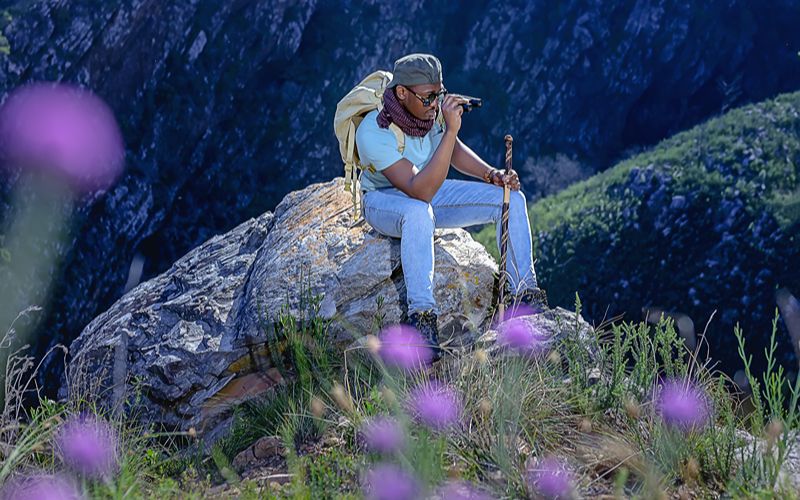 Eco cabins-a-young-african-male-hiking-in-the-mountains-amongst-pink-fynbos-flowers-during-spring-time-in-south-africa-865917817715-min